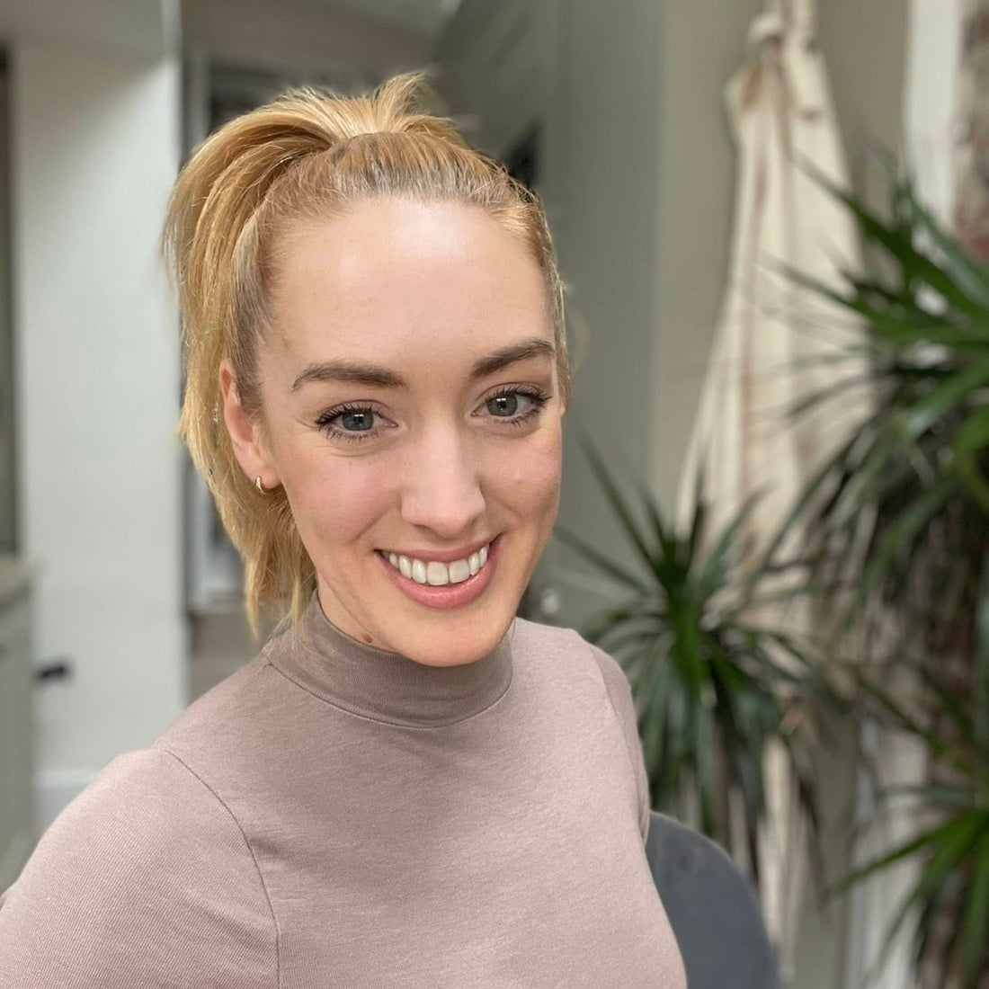 Woman in a beige turtleneck smiling outdoors with plants in the background